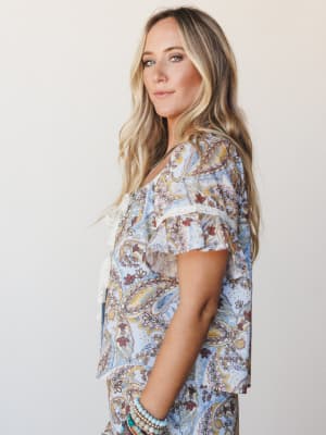 A soft side-profile shot of a model in a matching paisley printed set — powder blue with warm rust and gold tones — styled with stacked turquoise bracelets and a delicate lace-trim detail that gives the whole look a quietly romantic, boho-at-brunch e