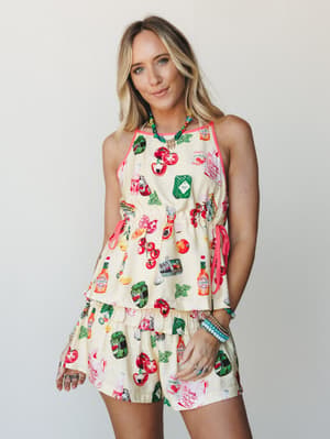 A playful matching short set in butter yellow covered in a cheeky food-themed novelty print — tomatoes, Tabasco bottles, pickles, herbs — styled with colorful beaded jewelry and a relaxed, sunny attitude.