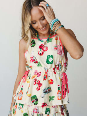 A smiling model in a playful butter yellow novelty-print set — covered in illustrated fruits, veggies, and Tabasco bottles — styled with stacked turquoise bracelets and a colorful statement necklace for a look that's equal parts whimsical and summer-