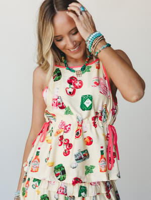 A smiling model in a playful butter yellow novelty-print set — covered in illustrated fruits, veggies, and Tabasco bottles — styled with stacked turquoise bracelets and a colorful statement necklace for a look that's equal parts whimsical and summer-