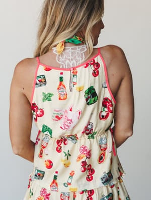 A back-view shot of a butter yellow short set covered in a playful illustrated food print — Tabasco bottles, tomatoes, cherries, and pantry staples — with coral piping trim, lace detail at the upper back, and a relaxed elastic waist that makes it fee