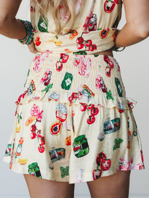 A close-up back shot of a matching short set in buttery cream, covered in a playful illustrated food print — tomatoes, jars, peppers, and produce — with a smocked waistband, ruffle hem shorts, and stacked beaded bracelets adding a relaxed boho touch.