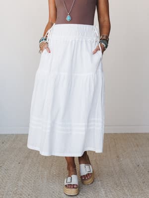 A white tiered maxi skirt with smocked waistband and side tie details, styled with a brown bodysuit, turquoise jewelry, and white platform espadrille sandals against a soft neutral interior backdrop.