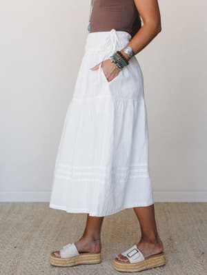 A breezy tiered midi skirt in off-white with tonal woven stripes, styled with a brown tank, stacked turquoise and beaded bracelets, and platform espadrille sandals — the kind of outfit that works equally hard at a farmers market or a summer dinner.