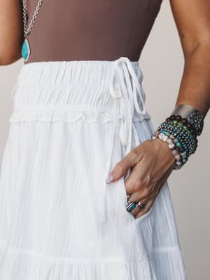A close-up waist shot of a white tiered maxi skirt with smocked waistband and drawstring tie, styled with a mocha bodysuit and layered boho jewelry — stacked beaded bracelets, a silver cuff, and a turquoise pendant necklace.