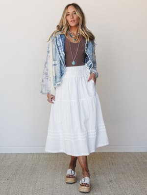A white tiered midi skirt styled with a distressed denim jacket, brown fitted tank, and platform espadrille sandals — layered with turquoise and beaded necklaces for that effortless boho-meets-everyday energy.