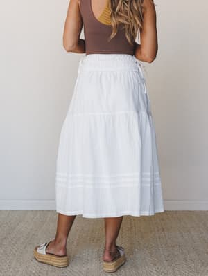 Shot from the back, a white tiered midi skirt with subtle woven texture pairs with a chocolate brown tank and platform espadrilles — easy, sun-warmed, and going somewhere good.