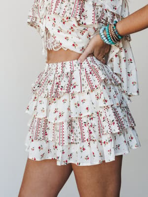 A flirty tiered ruffle skort in a cream ditsy floral print styled with a matching crop top and stacked turquoise bracelets — the kind of outfit that makes a Tuesday feel like a road trip.