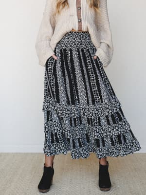 A tiered maxi skirt in a black and white mixed floral stripe print is styled with a cream chunky knit cardigan and black ankle boots — the kind of outfit that looks like it came together effortlessly but was definitely thought about.