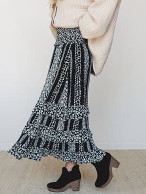 A black and white floral-stripe maxi skirt with tiered ruffles and a smocked waist, styled with a cream chunky knit sweater and black suede wedge ankle boots — the kind of outfit that works just as hard at a weekend market as it does at a fall dinner