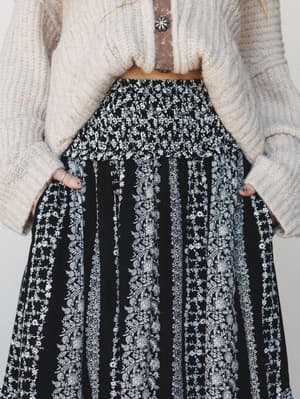 A close-up torso shot of a flowy black maxi skirt covered in delicate white floral stripe print, styled with a cropped cream knit sweater and a brown leather strap with a silver concho detail — the kind of outfit that looks effortless but was definit
