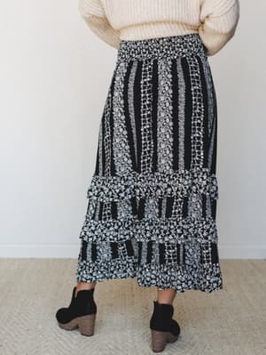 Shot from behind, this black floral midi skirt pairs with a cream chunky knit sweater and black ankle booties — the kind of fall outfit that feels pulled-together without trying too hard.