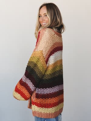 A model glances back over her shoulder with an easy smile, wearing an oversized multicolor striped knit cardigan in warm autumn tones — the kind of piece that looks like it was pulled from a very well-loved closet.