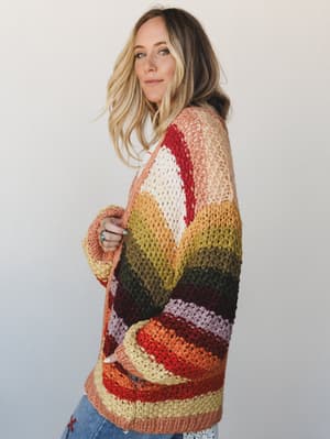 A model shot in soft studio light wearing an oversized, chunky crochet-knit cardigan in a warm autumn stripe palette — the kind of sweater that looks like it was made for wandering farmers markets and long porch mornings.