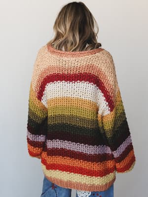 Shot from behind, a model wears an oversized crochet cardigan in a full spectrum of autumn-toned stripes — peachy blush to deep olive to burnt orange — layered over jeans, giving off that effortlessly cozy, color-happy energy.
