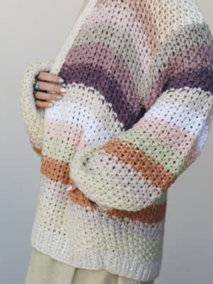 A close-up of a chunky knit cardigan in soft multicolor stripes — cream, blush, mauve, purple, sage, and camel — worn with arms crossed, showing off stacked rings and painted nails that feel like part of the outfit.