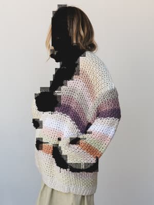 A model shot from behind in a chunky knit cardigan with soft multicolor stripe blocking — cream, blush, lavender, sage, and terracotta — worn over what looks like a relaxed olive skirt, photographed against a clean light gray studio backdrop.