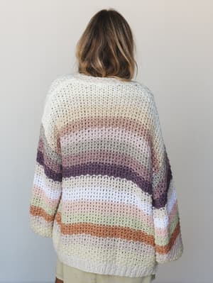 Shot from the back, a model wears an oversized crochet-knit cardigan in a soft, dreamy stripe sequence — cream fading into blush, sage, plum, and terracotta — the kind of sweater that looks like it was made for curling up somewhere cozy.