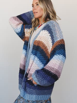 A chunky multicolor striped crochet cardigan worn oversized and relaxed, layered over a white tee with a beaded statement necklace — the kind of sweater you reach for every single time.