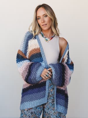 A chunky striped knit cardigan in shades of blue, mauve, and warm brown layered over a white tank and butterfly-print pants — the kind of outfit that feels like a soft landing after a long week.