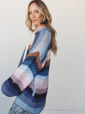 A model looks back over her shoulder wearing an oversized multicolor stripe knit cardigan — the kind of piece that looks like it took all weekend to make and feels like a hug when you put it on.
