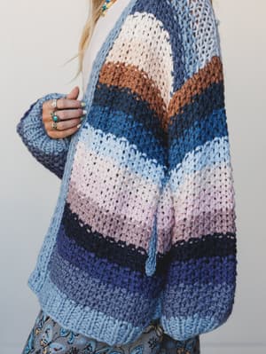 A close-up of a chunky crochet cardigan in a dreamy stripe of slate blue, dusty rose, caramel, and navy — stacked rings and a peek of printed pants underneath make it feel effortlessly collected.