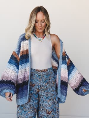 A relaxed, dreamy look built around an oversized chunky crochet cardigan in multicolor stripes — worn open over a white tank and butterfly-print wide-leg pants, layered with colorful beaded necklaces for that effortlessly expressive, boho-at-home ene