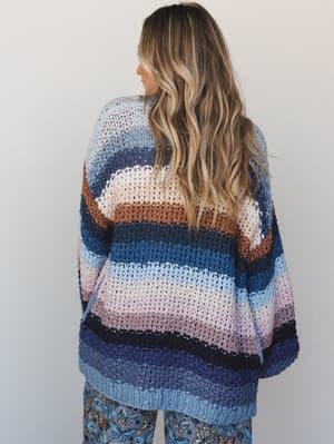 A back-facing model shows off a chunky open-knit striped cardigan in a dreamy mix of blues, lavender, blush, and caramel, paired with a butterfly-print pant — the kind of outfit that looks like it was assembled by someone who just *gets it*.