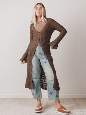 Woman wearing a long brown cardigan over a patterned dress with light blue jeans against a plain background