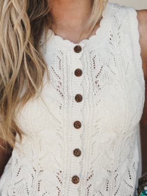 A close-up torso shot of a cream pointelle knit sleeveless vest with wooden button-front closure and delicate scalloped neckline — the kind of piece that looks just as good over a sundress as it does with cutoffs.