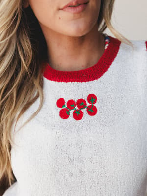 A close-up of a cream knit sweater with a bold red ribbed neckline and a cheerful cluster of cherry tomato embroidery at the chest — the kind of piece that makes you smile before you've even put it on.