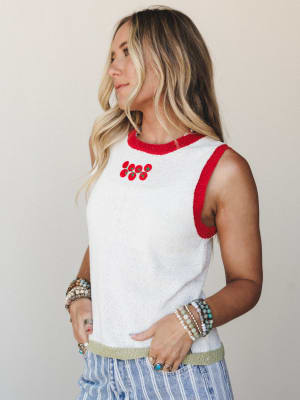 A sun-kissed model wears a cropped knit tank in off white with bold red ringer trim and a small tomato embroidery cluster at the chest, layered over striped wide-leg pants and stacked with beaded and stone bracelets — casual and full of personality.