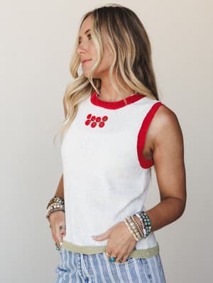 A sun-kissed model wears a cropped knit tank in off white with bold red ringer trim and a small tomato embroidery cluster at the chest, layered over striped wide-leg pants and stacked with beaded and stone bracelets — casual and full of personality.