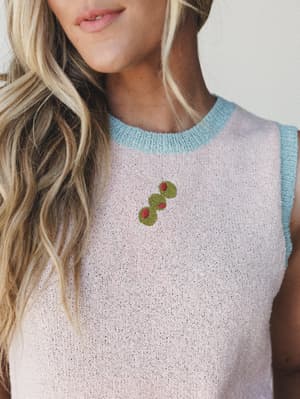 A close-up of a blush knit sleeveless tank with a soft blue contrast neckline and armhole trim, featuring a small embroidered cactus detail at the chest — the kind of top that makes you feel put-together without trying too hard.