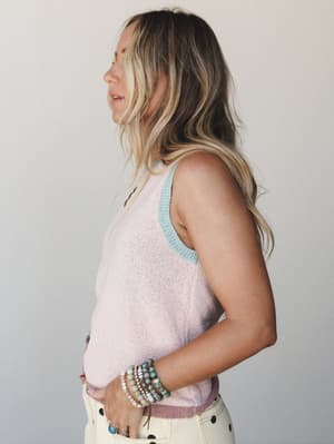 A side-profile shot of a model in a soft pink knit tank with mint contrast trim, layered beaded bracelets, and cream polka dot pants — casual and collected with a quietly personal feel.