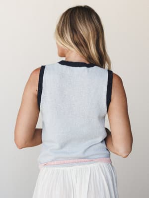 Shot from the back, a model wears a pale blue sleeveless knit tank with navy contrast trim at the neckline and armholes and a soft pink ribbed hem, paired with a white gauzy midi skirt — easy, pretty, and just the right amount of put-together.