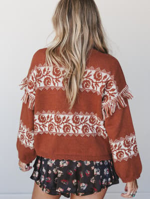 Shot from behind, a model layers a rust and cream fair isle knit sweater with fringe shoulder trim over a dark floral mini skirt — cozy, boho, and very much the outfit you wear when the season finally tips into something cooler.