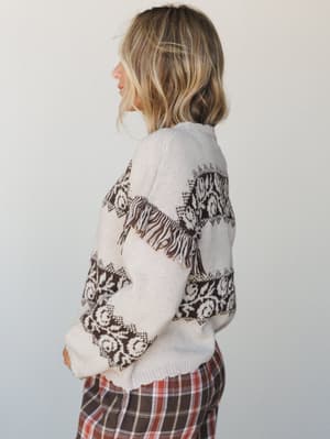 A side-profile shot of a model in a cream knit sweater with dark brown floral and scrollwork jacquard patterning and fringe trim at the waist, paired with rust and brown plaid wide-leg pants — the kind of outfit that feels like autumn finally showed