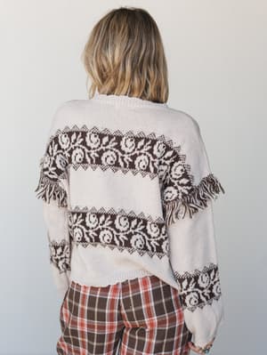 Shot from the back, this cream knit sweater with chocolate brown floral fair isle bands and playful fringe trim is paired with rust plaid wide-leg pants — the kind of cozy outfit that feels like autumn in the best possible way.