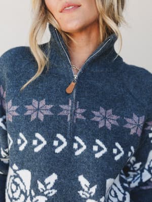 A close-up detail shot of a cozy quarter-zip sweater in deep indigo, featuring a fair isle-inspired snowflake and floral knit pattern in cream and dusty mauve, finished with a warm tan leather zipper pull.
