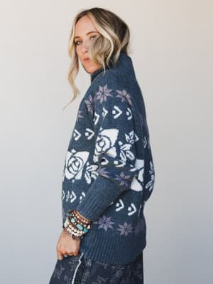 A model shot from the side showing a cozy oversized turtleneck sweater in deep indigo with a rose and snowflake Fair Isle knit pattern, styled with printed wide-leg pants and a stacked bracelet set — the kind of sweater that makes a Tuesday feel inte