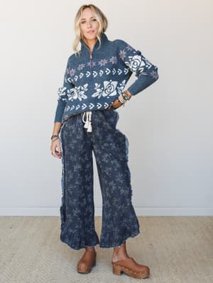 A full-body studio shot of a model wearing a cozy navy fair isle sweater with rose and snowflake intarsia paired with dark floral wide-leg pants with ruffle hems and frayed seams, finished with brown platform clogs — the whole look feels like a fall