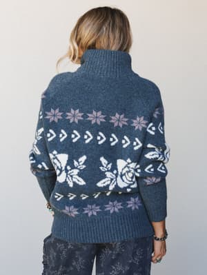 A back-view shot of a cozy fair isle turtleneck sweater in deep indigo, featuring a mix of rose, snowflake, and geometric intarsia knit patterns — the kind of sweater that makes you want to pour something warm and stay a while.