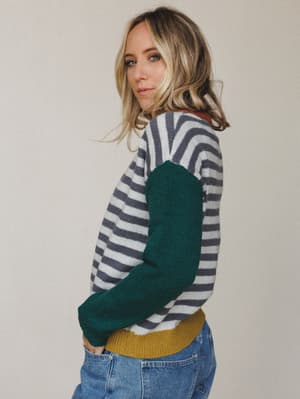 A model shot over the shoulder in a colorblock striped knit sweater — grey and cream stripes across the body, a deep forest green sleeve, and a mustard ribbed hem that makes the whole thing feel like a cozy Saturday with good taste.