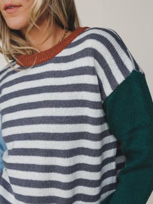 A close-up of a cozy knit sweater with classic grey and white stripes, a rust-colored ribbed neckline, and deep teal colorblock sleeves — the kind of sweater that makes a Tuesday feel intentional.