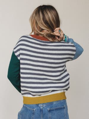 Shot from the back, she's holding her hair up to show off a colorblock striped sweater — grey and cream stripes across the body, teal left sleeve, dusty blue right sleeve, rust neckline, and a mustard ribbed hem, paired with light wash jeans and a st