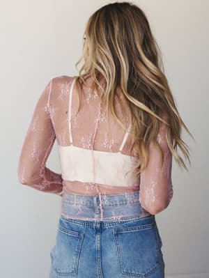 Shot from behind, a model layers a sheer blush lace long-sleeve top over a cream bralette, paired with light-wash denim — the kind of effortlessly romantic combination that looks like it came together without trying.
