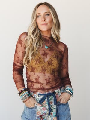 A sheer floral lace long-sleeve top in deep burgundy layered over a mustard crochet bralette, styled with floral-patch jeans and a stack of beaded bracelets — the kind of outfit that looks like it came together effortlessly but definitely didn't.