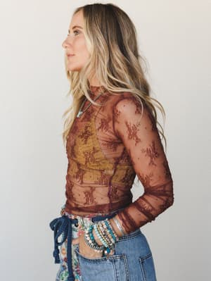 A side-profile shot of a model in a sheer rust-toned lace top layered over a mustard cami, paired with light-wash jeans and stacked beaded bracelets — the kind of outfit that looks like it came together effortlessly but absolutely did not.