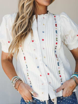 A white embroidered blouse with scattered multicolor floral stitching and lace trim details, styled with stacked beaded bracelets and polka dot jeans — sweet, lived-in, and ready for a sun-filled afternoon.